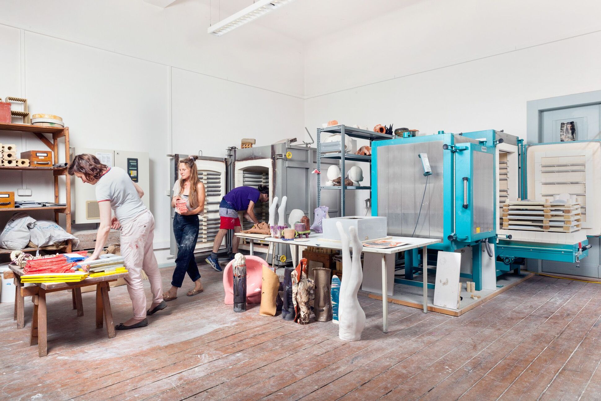 Keramikwerkstatt. Verschiedene Skulpturen, Gipsformen, diverse Oberflächenbehandlungen sowie Glasuren und Engoben, Brennofen für Keramik und Ton und andere Objekte im Raum. Drei Studenten beim Arbeiten.