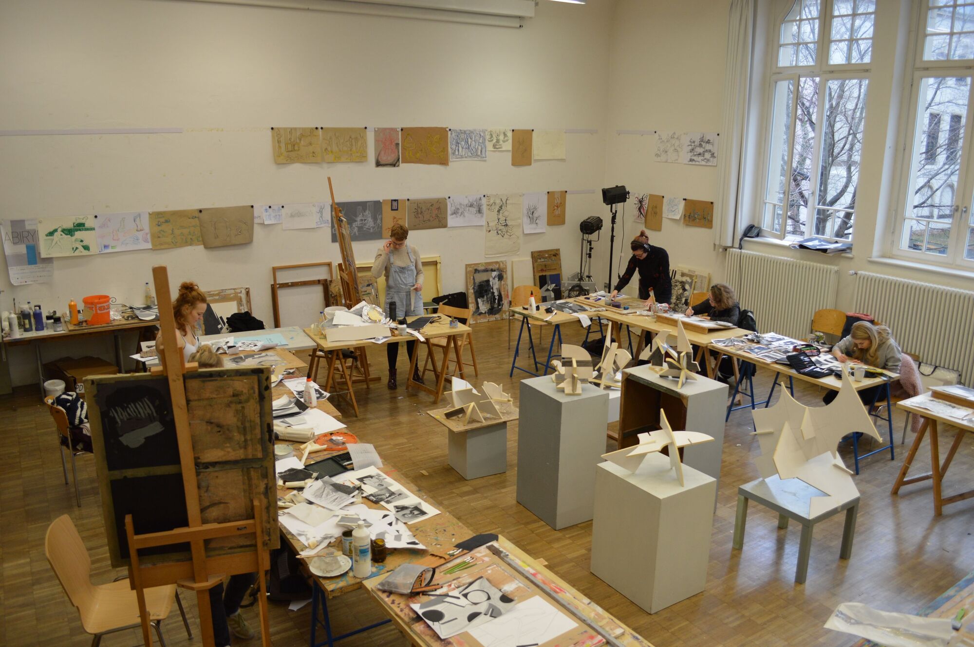 In der Mitte des Ateliers ein Stillleben bestehend aus mehreren geometrischen Objekten. Um das Stilleben herum stehen Tische. Studenten beim Schaffen.