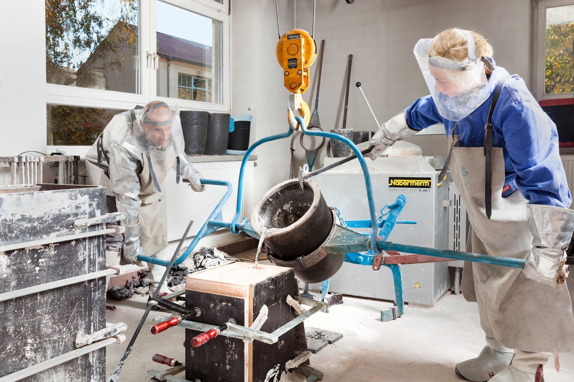 Zwei Menschen in entsprechender Bekleidung beim Gießen von Gusseisen in eine Sandform.