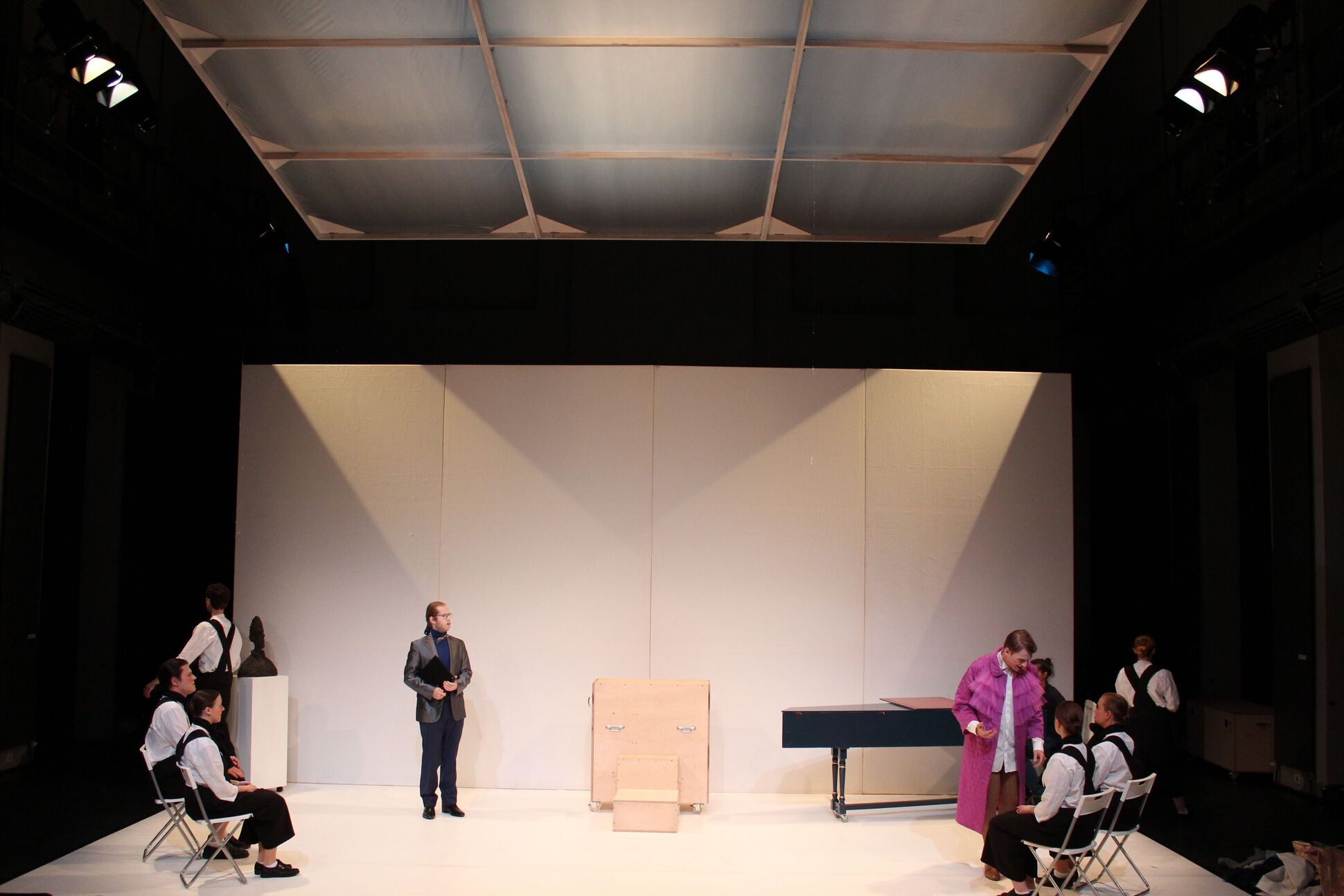 Bühnenbild: Auf der weißen Bühne sind Menschen und verschiedene Objekte zu sehen. Bühnenbild: Auf der weißen Bühne sind Menschen und verschiedene Objekte zu sehen.