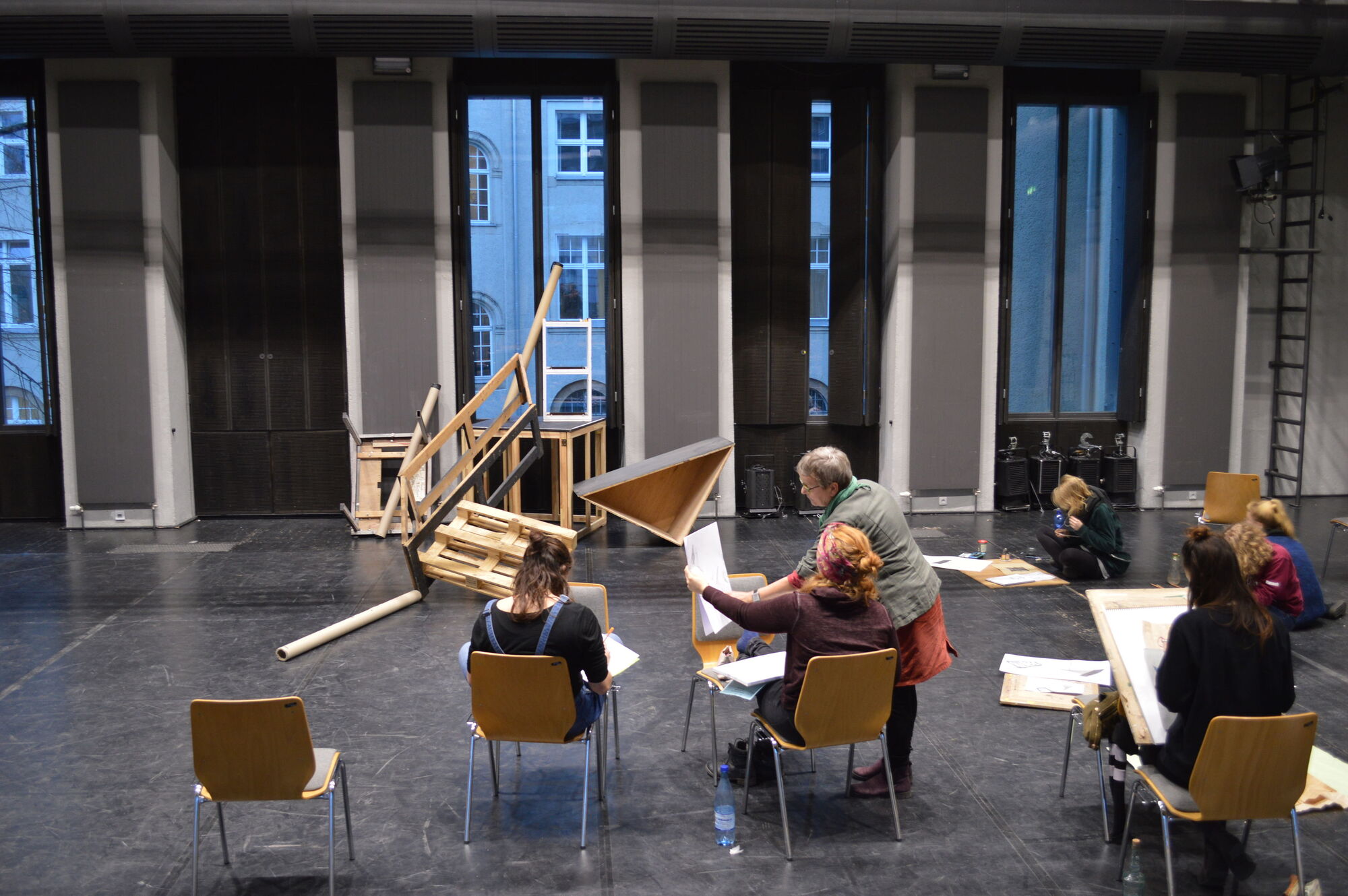 Im Hintergrund ein Stillleben bestehend aus großen geometrischen Objekten. Im Vordergrund Studenten beim Schaffen.