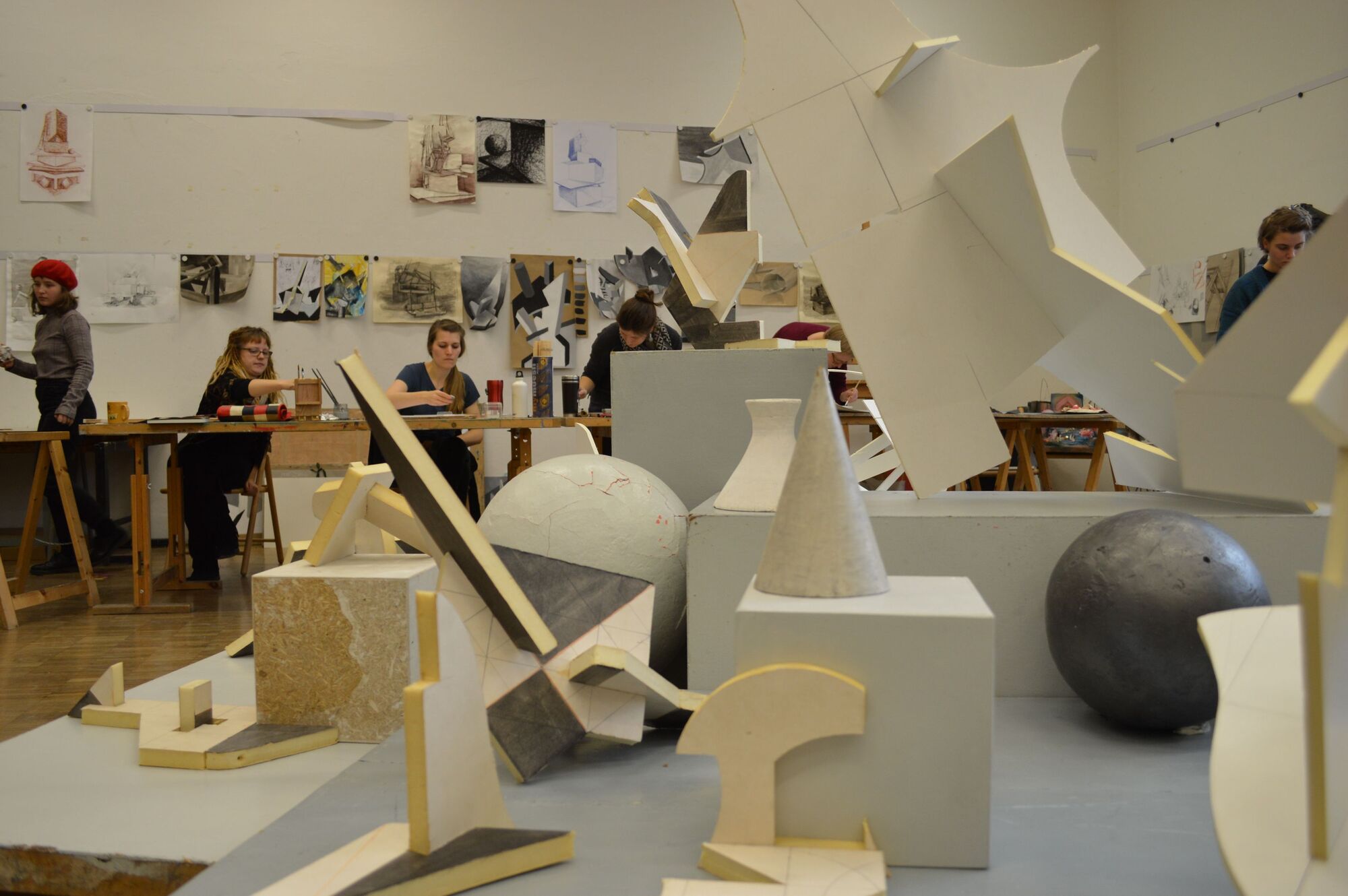 Im Vordergrund ein Stillleben bestehend aus mehreren geometrischen Objekten. Im Hintergrund Studenten beim Schaffen.