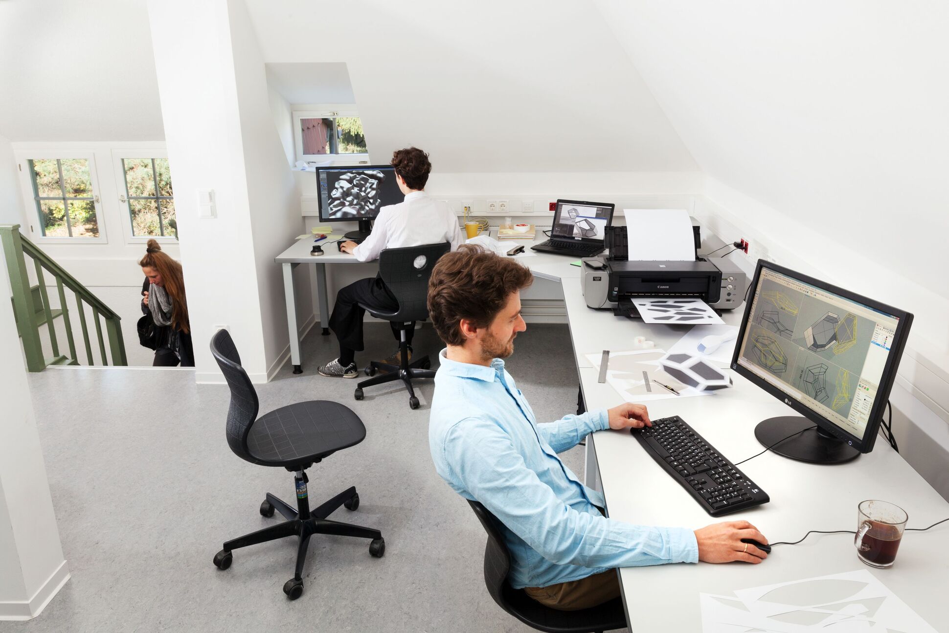 Zwei Personen sitzen am Computer. Auf dem Tisch sind Kopiermaschine zu sehen. Studentin im Hintergrund.