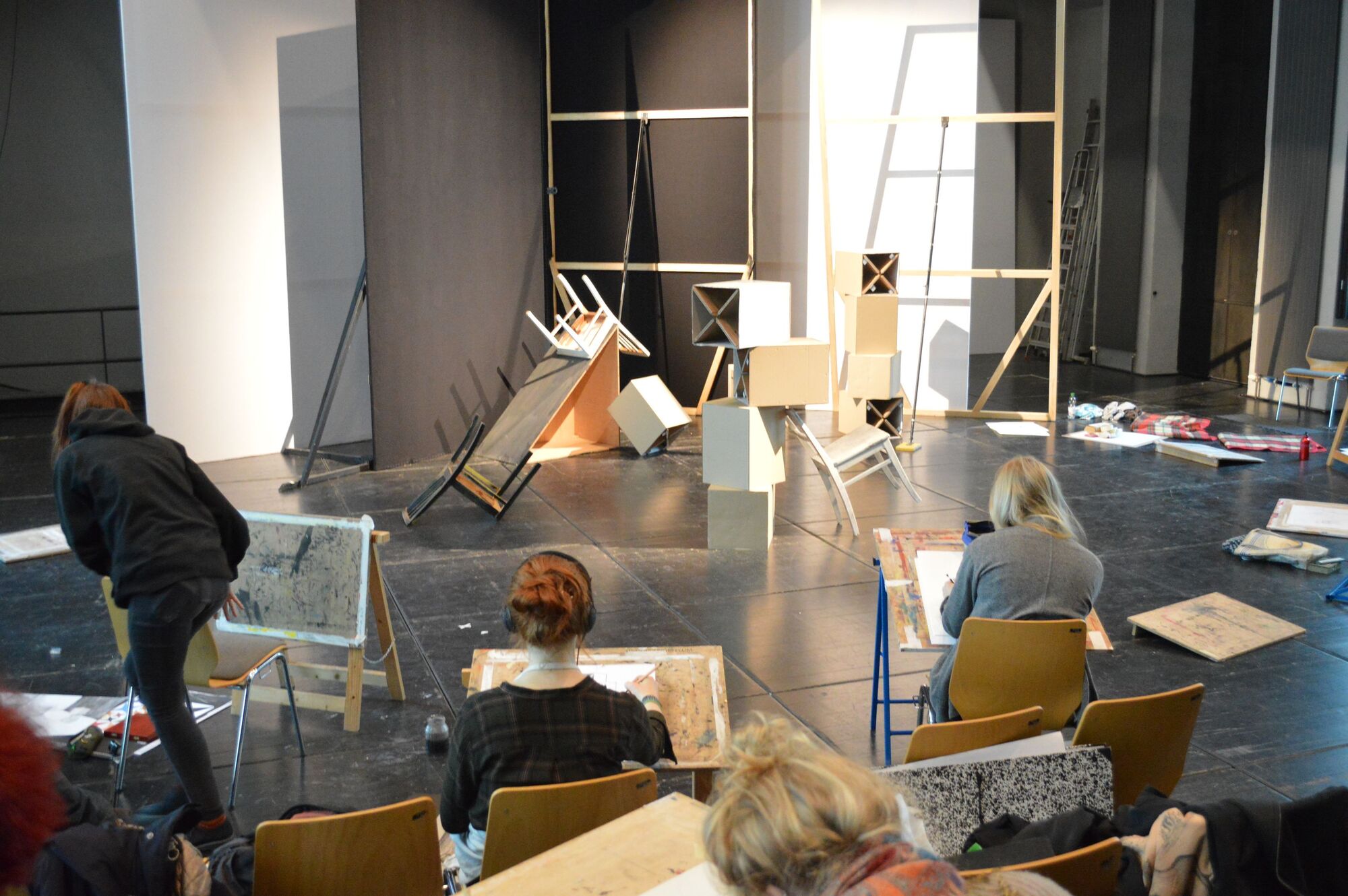Im Hintergrund ein Stillleben bestehend aus großen geometrischen Objekten. Im Vordergrund Studenten beim Schaffen.