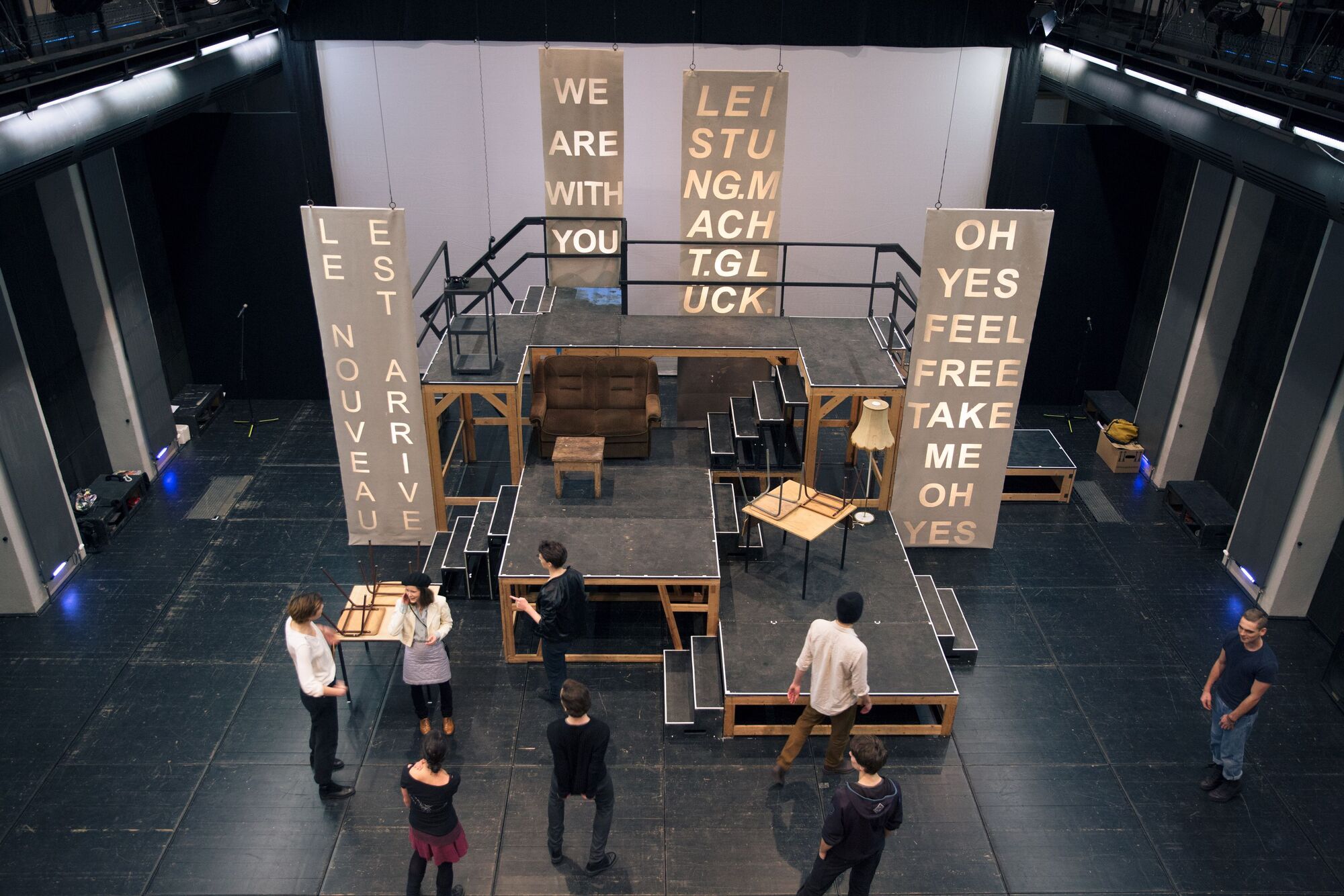 Bühnenbild: Menschen und verschiedene Möbel auf der Bühne. 