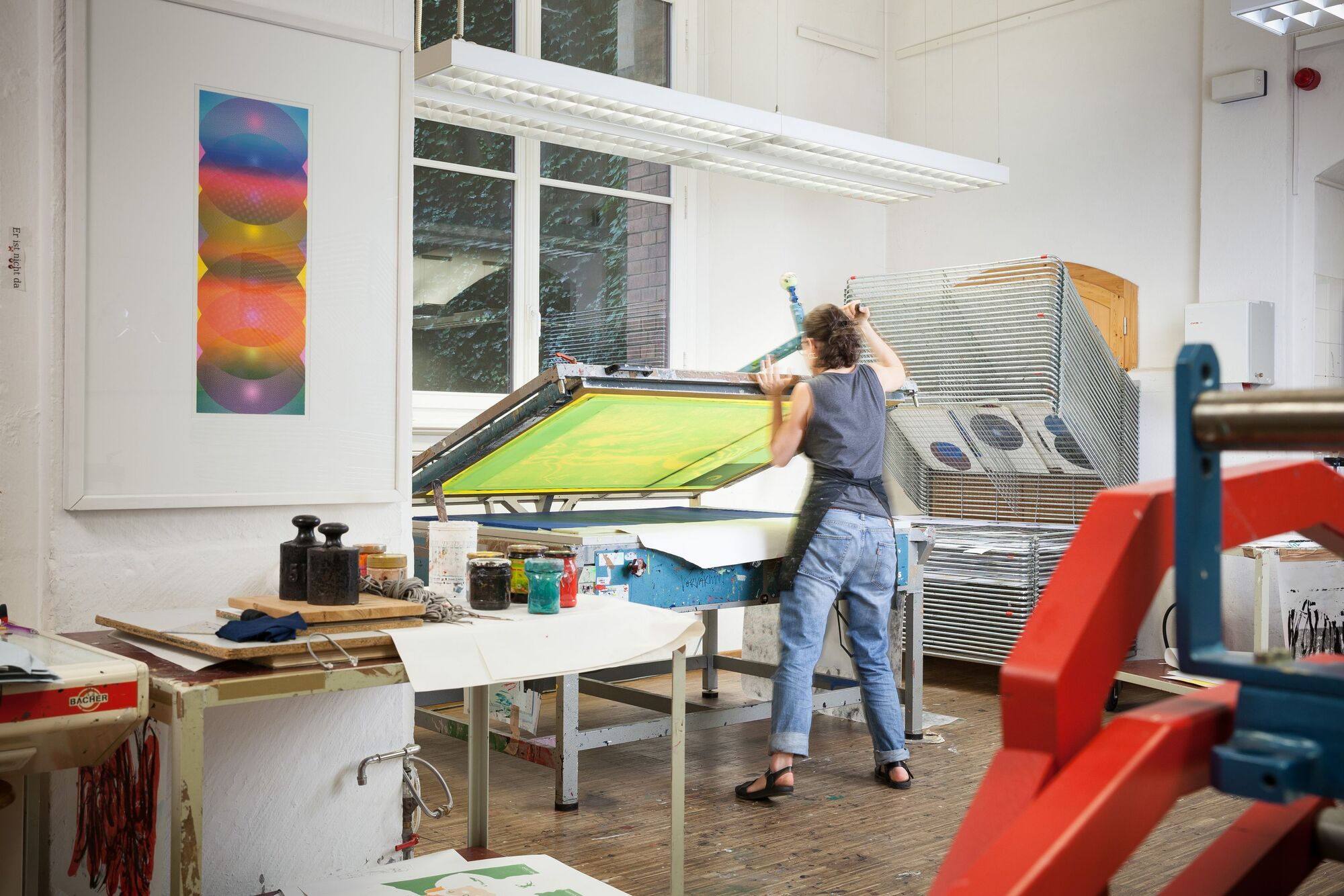 Werkstatt für Siebdruck. Siebdruckpresse, Stapeltrockner, Siebdruckplatten, verschiedene Farben und andere Werkzeuge für Siebdruck. Eine Studentin beim Arbeiten.