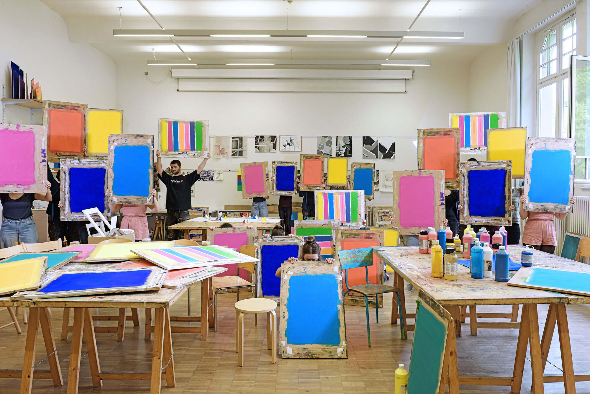 Atelier des Studienganges künstlerische Grundlagen. Tische, Stühle, Staffeleien und Farben im Raum. Bilder auf der Wand. Jeder Student der Fachklasse hält ein einfarbiges Bild. 