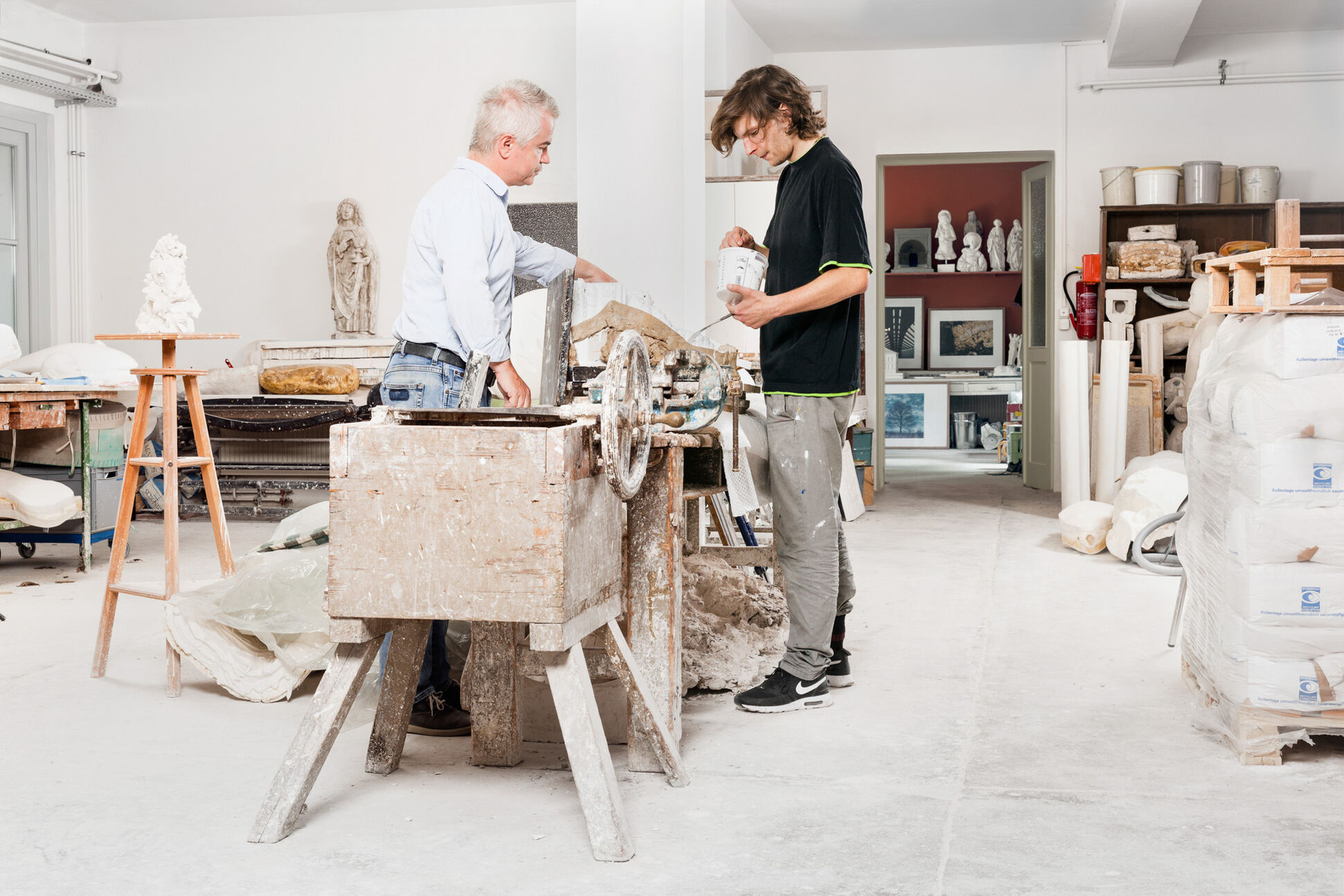 Zwei Personen machen eine Gipsform in der Werkstatt für Abformungen. In dem Raum sind Skulpturen, Staffeleien, Plastikbecher und andere Objekte zu sehen.