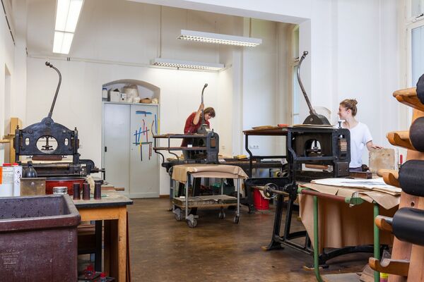 Werkstatt für Lithografie. Lithografiesteine, Steinhandhebelpressen, Papiere, Regale, Arbeitstische und andere Objekte. Zwei Studentinnen beim Arbeiten.