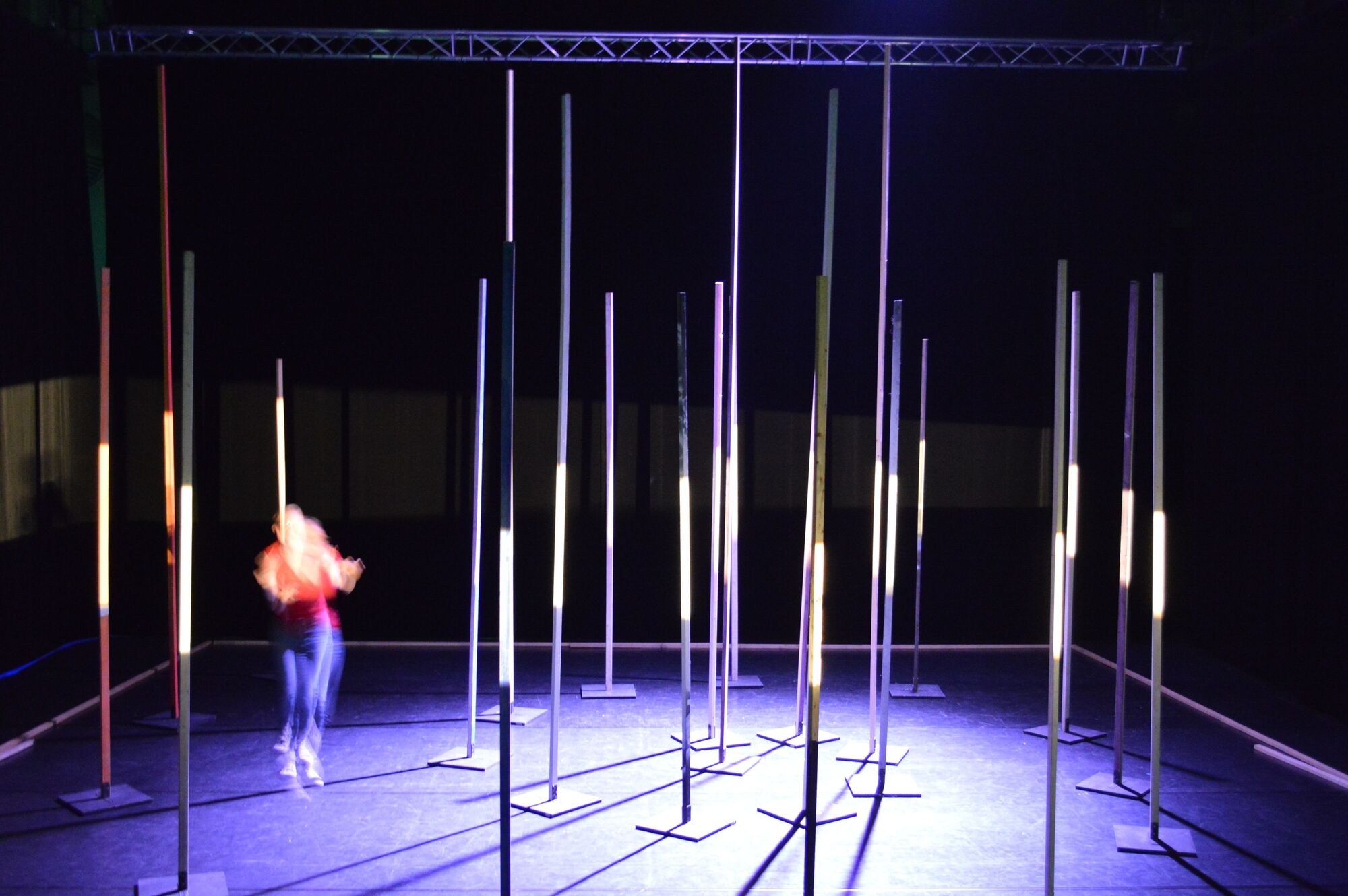 Vertikale Holzlatten leuchten in lila auf der Bühne. Eine Studentin führt zwischen den Holzlatten einen Performance durch.