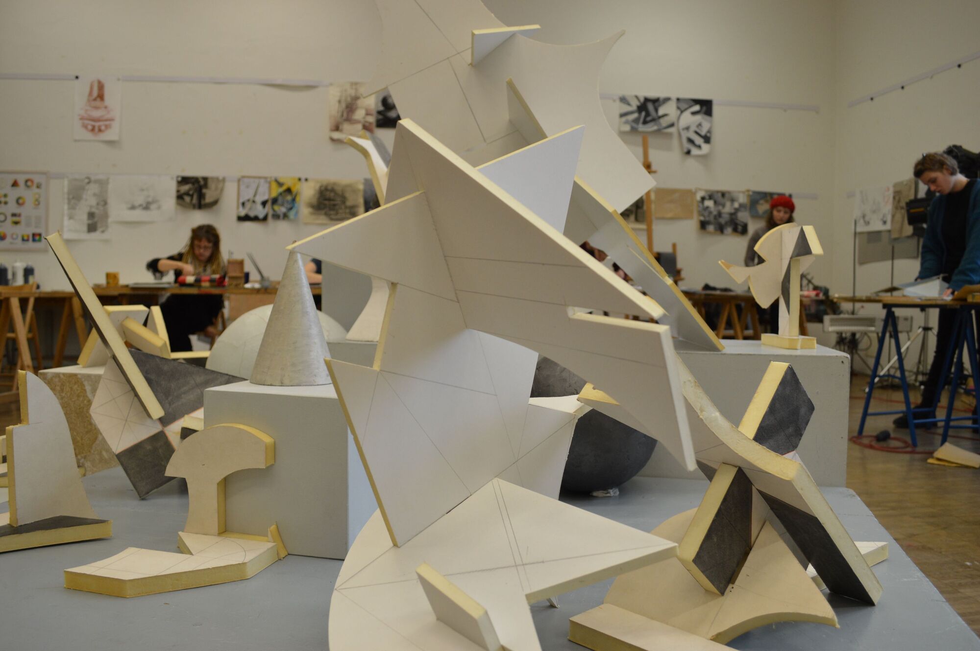 Im Vordergrund ein Stillleben bestehend aus mehreren geometrischen Objekten. Im Hintergrund Studenten beim Schaffen.