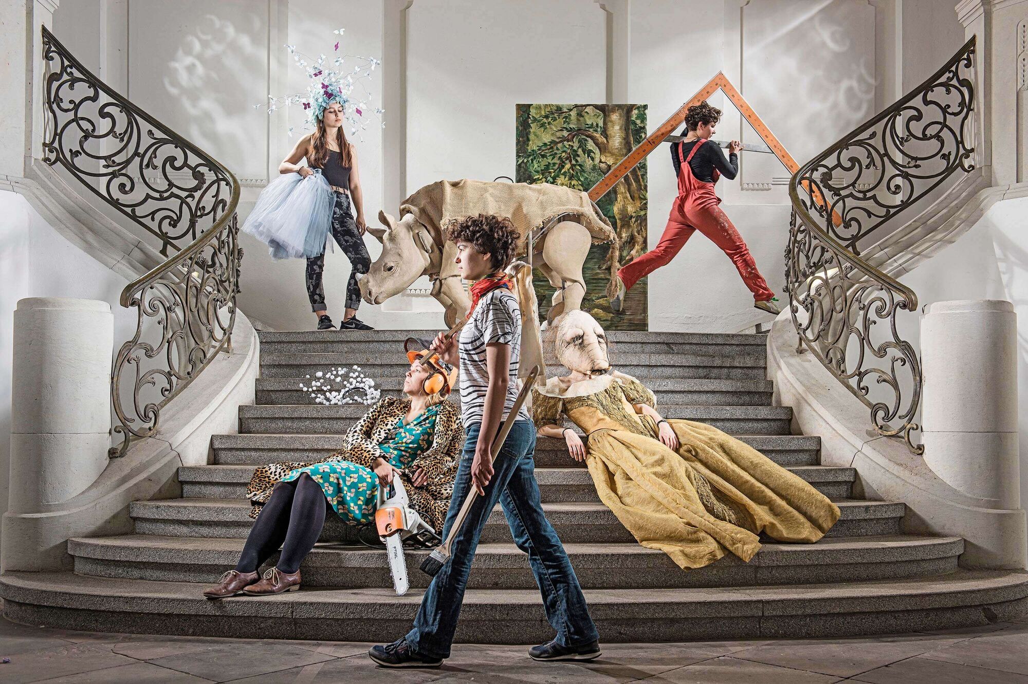 Breite Treppe in der Flur der Hochschule. Zwei Studentinnen in bunten Kostümen sitzen an der Treppe. Weitere drei Studentinnen in verschiedenen Kostümen laufen auf der Treppe. In der Mitte des Bildes, im Hintergrund, sieht man eine Nashorn-Figur und ein Gemälde in grünen Farbtönen.