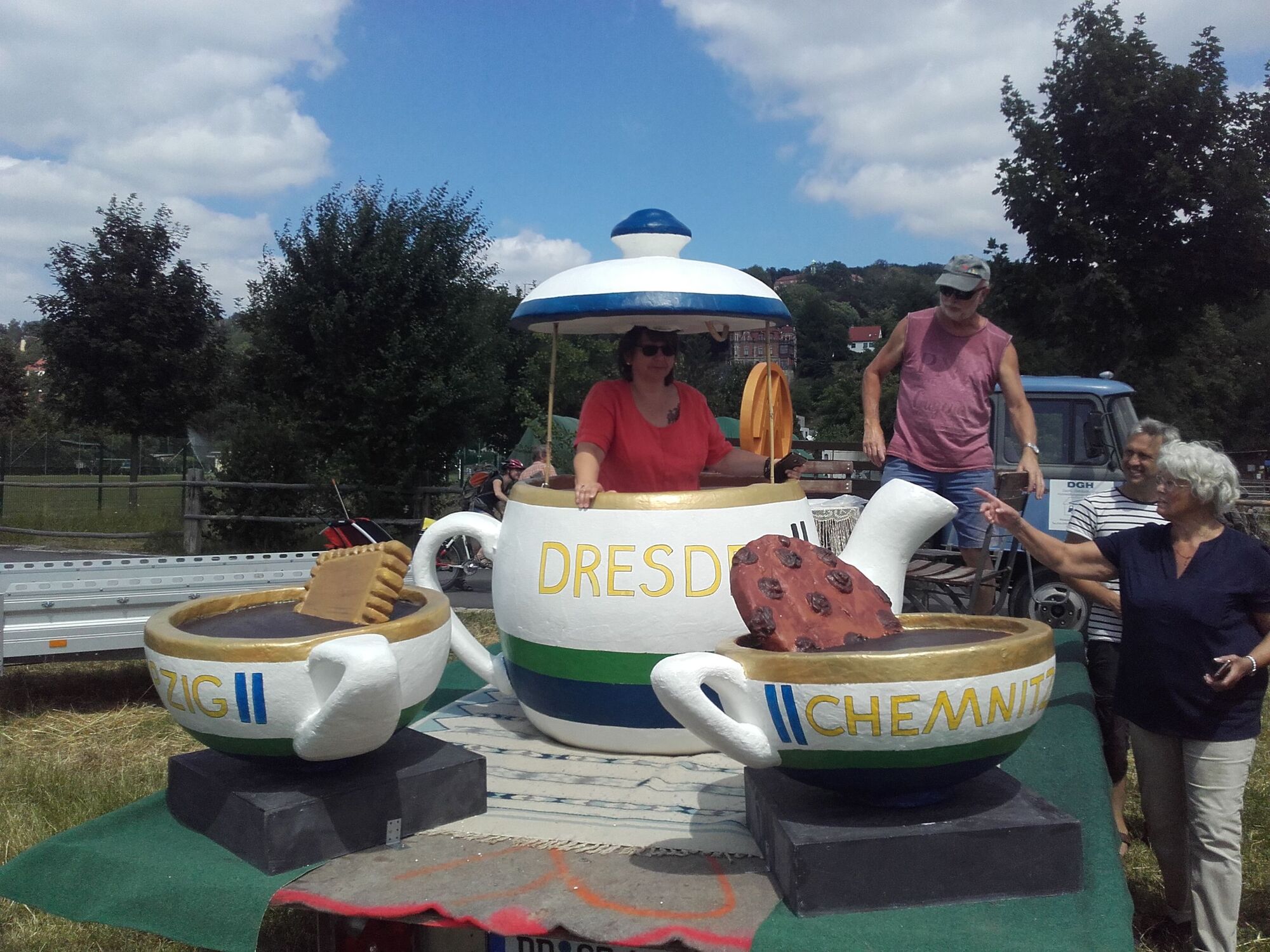 Menschen gucken eine lebensgroße Tee-Set an. Eine Frau sitzt in der lenensgroßen Teekanne.