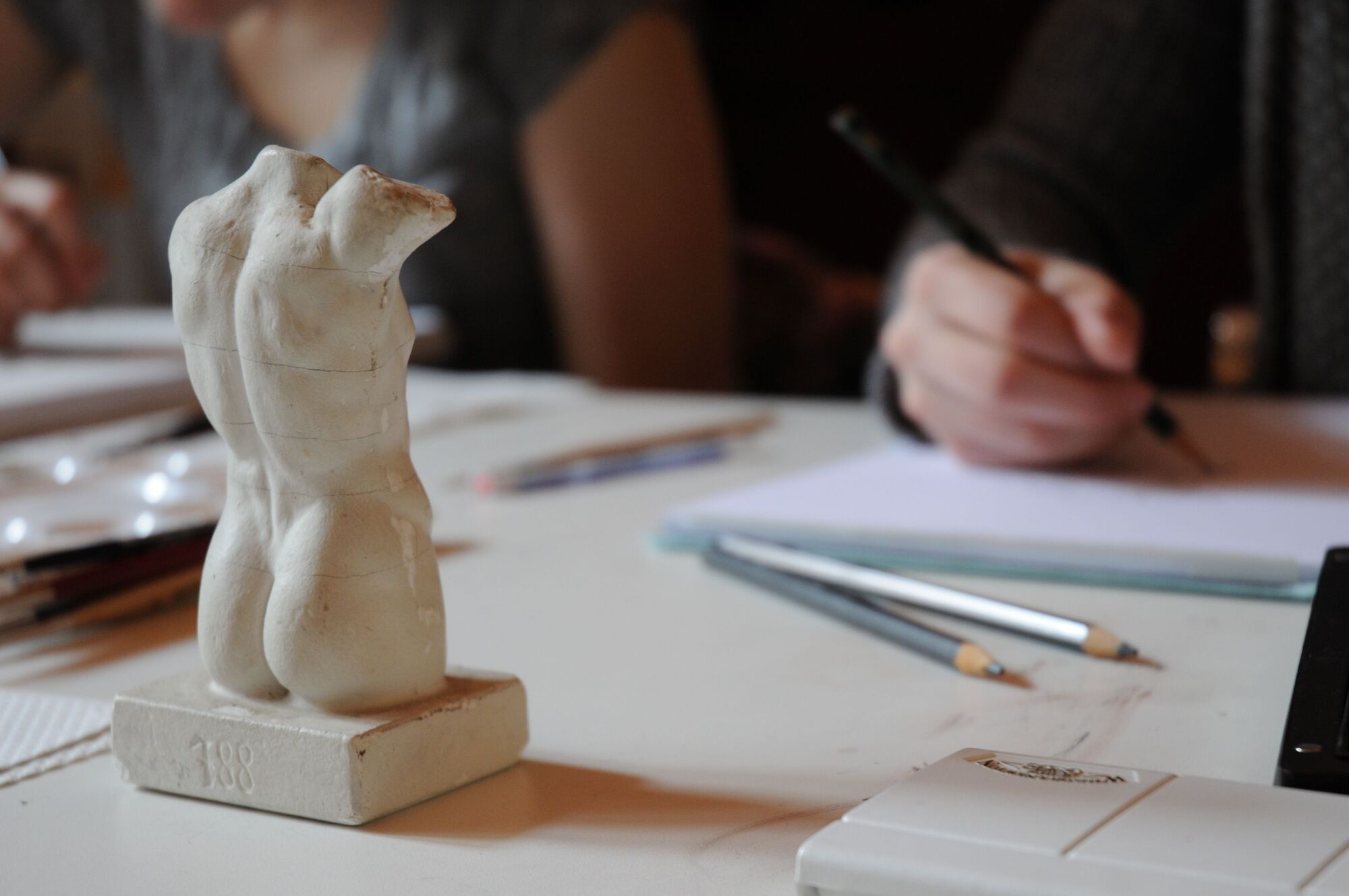 Im Vordergrund ein kleiner Torso aus Stein. Studentin zeichet ihn mit einem Zeichenstift.