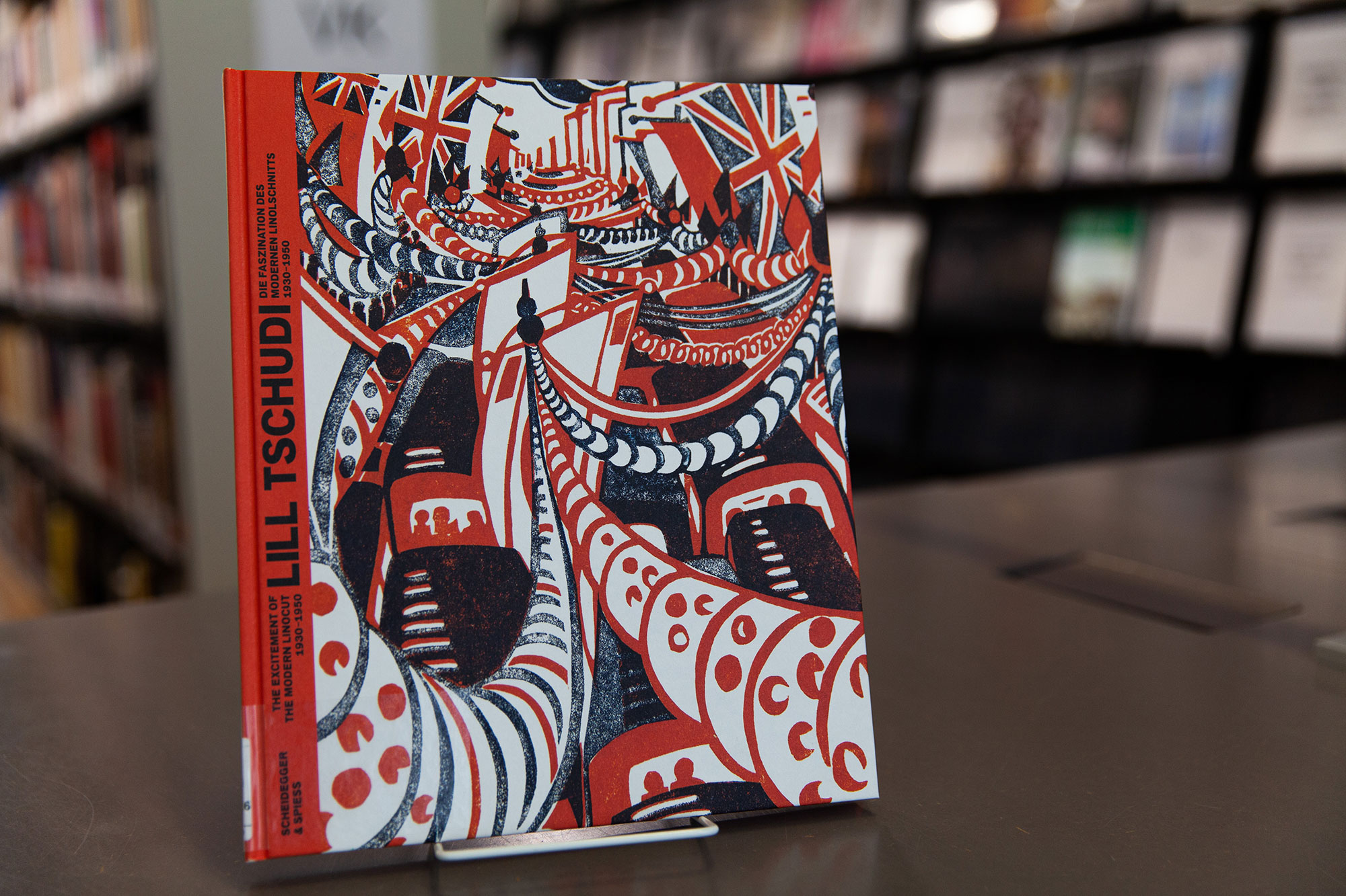 Ansicht des Buchs "Lill Tschudi: die Faszination des modernen Linolschnitts", aufgestellt, im Hintergrund Bibliotheksregale