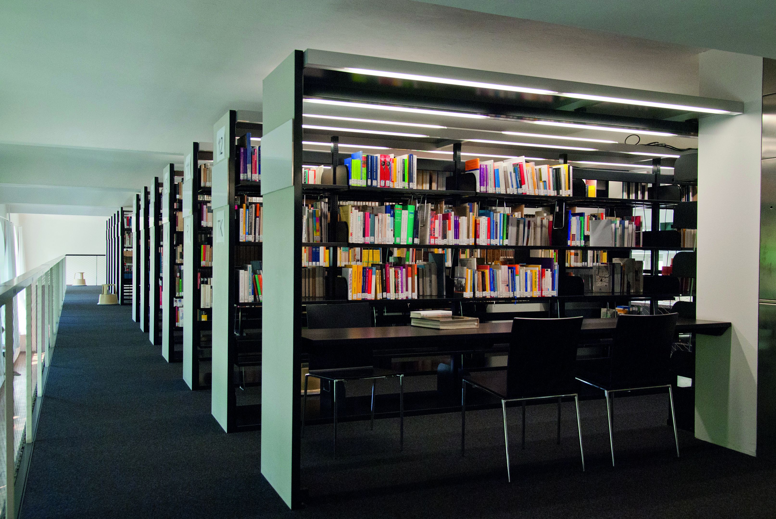 Bibliothek der Hochschule. Mehrere Bücherregale. Schwarzer Tisch mit Stühlen.