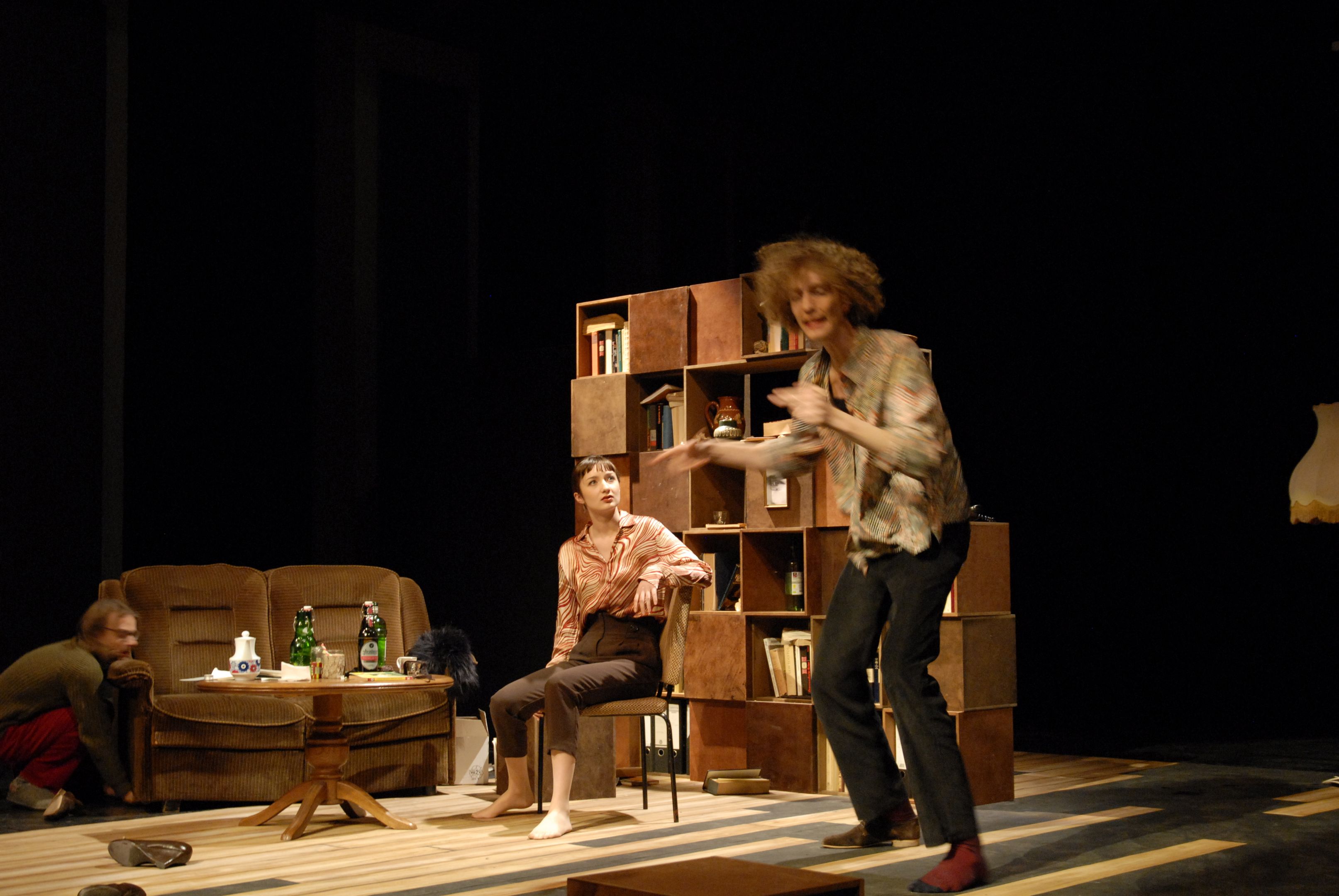 Bühnenbild: Drei Menschen im Wohnzimmer. Bücherregal, Sofa, Wohnzimmertisch und andere Objekte.