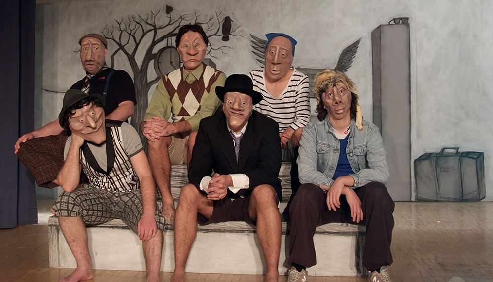 Szenenfoto, eine Gruppe von sechs Personen mit Masken sitzt auf einem Podest