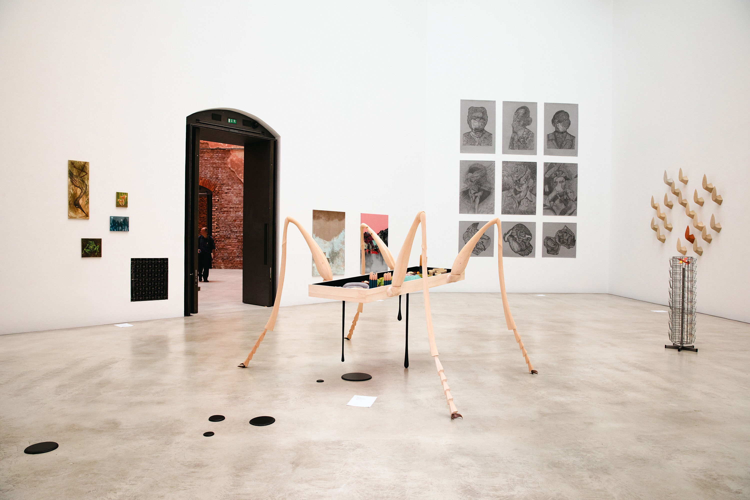 Ausstellungsansicht: Bilder an der Wand und eine große Holzskulptur.