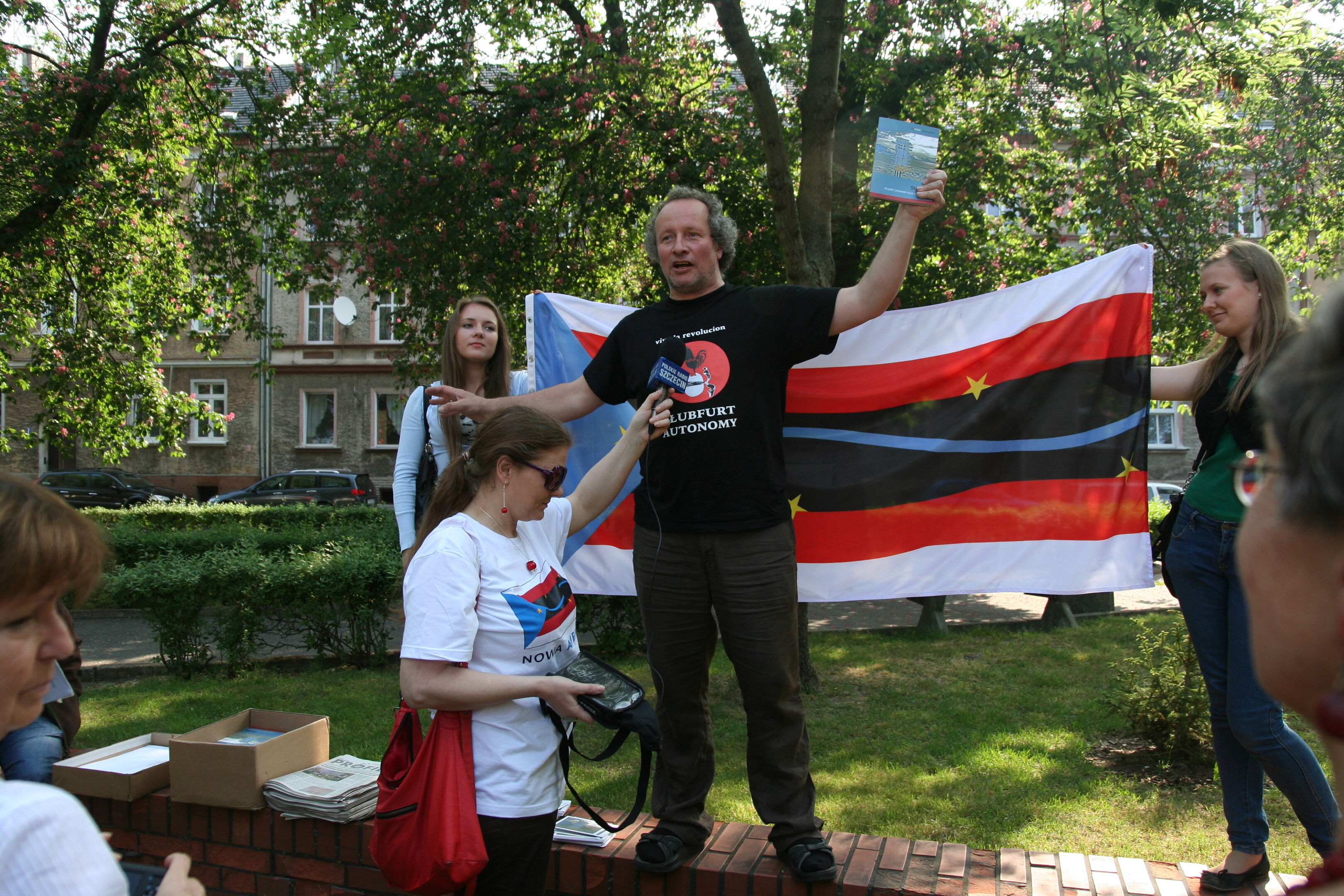 Eine männlich gelesene Person steht mit einem Buch in der Hand auf einer städtischen Grünfläche.